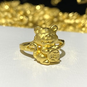 Könnte beinhalten: Ein goldener Ring mit einem Cartoon-Tiger, der einen runden Anhänger hält. Der Ring ist ein traditionelles chinesisches Design.