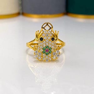 May include: A gold-toned ring shaped like a frog, featuring a body encrusted with clear white stones, two black stone eyes, and a central flower motif composed of green stones surrounding a single pink stone at the center.