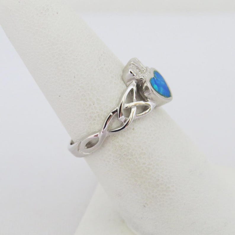 K&ouml;nnte beinhalten: Ein silberner Ring mit einem blauen Opal-Herz und einem keltischen Knotendesign.