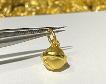 May include: A gold-colored bell charm pendant with a small loop at the top. The charm is held by a pair of tweezers.