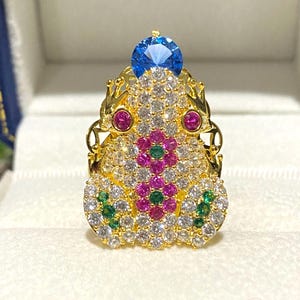 以下が含まれることがあります： 青い宝石が上にあり、透明な宝石とピンクの宝石で囲まれた金の蛙の指輪。蛙の目は赤い宝石で、体は緑の宝石で飾られています。