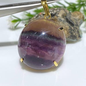 May include: A gold pendant featuring a large, oval-shaped fluorite gemstone with purple, green, and pink hues. The gemstone is set in a gold bezel and has a smooth, polished finish.