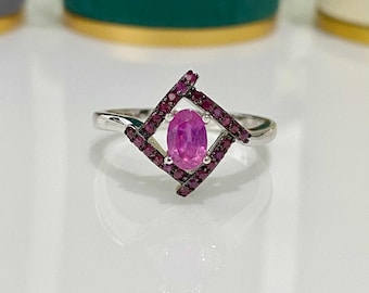 Könnte beinhalten: Ein silberner Ring mit einem ovalen, rosafarbenen Edelstein in der Mitte, eingerahmt von einem geometrischen Design aus kleineren, dunkelrosa Steinen. Der Ring hat ein poliertes Silberband. Das Design ist modern und auffällig.