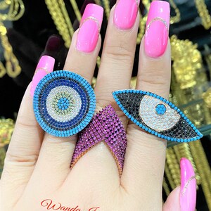 May include: A hand displays four rings: a round blue and white evil eye, a triangular pink ring, and an eye-shaped ring with black and turquoise accents. The nails are painted bright pink with gold accents. The text "Wanda Jewelry Etsy" is visible.
