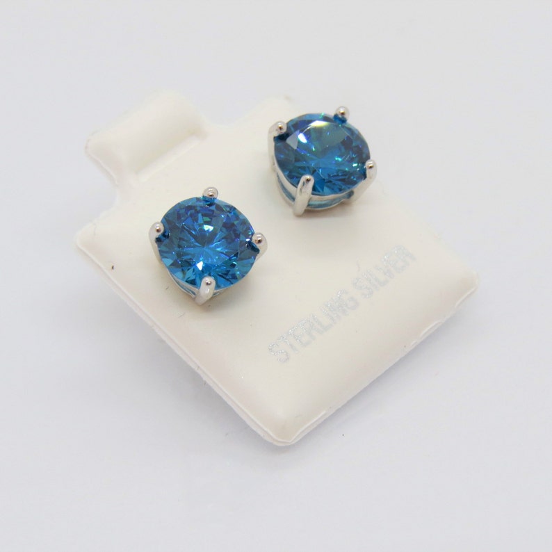 Puede incluir: Un par de pendientes de plata de ley con piedras redondas azules. Los pendientes se muestran en una tarjeta blanca con las palabras "Sterling Silver" impresas en ella.