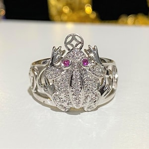 May include: A silver ring with a detailed frog design. The frog is covered in small white stones and has two pink gemstones for eyes. The ring band is also decorated with a silver design.
