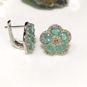 May include: A pair of silver-toned earrings. One earring features a flower-shaped design with five oval-cut emeralds surrounded by small pink and yellow gemstones. The other earring has a rectangular shape with multiple emeralds. The earrings are set against a white background.