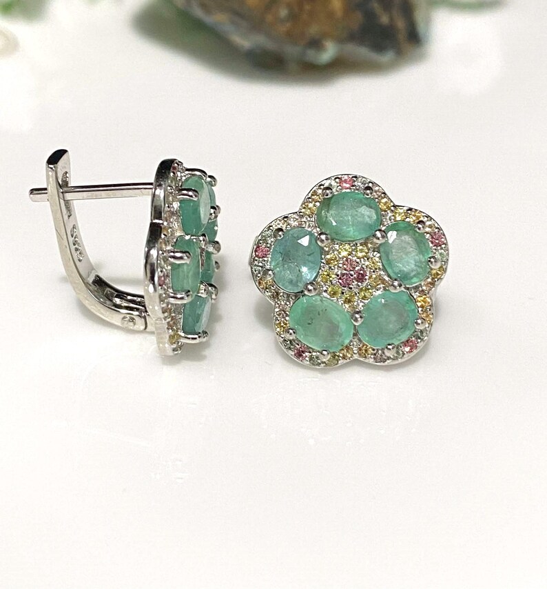 May include: A pair of silver-toned earrings. One earring features a flower-shaped design with five oval-cut emeralds surrounded by small pink and yellow gemstones. The other earring has a rectangular shape with multiple emeralds. The earrings are set against a white background.