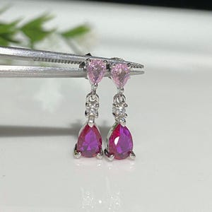 Puede incluir: Un par de pendientes colgantes con piedras preciosas en forma de pera rosa y roja. Los pendientes son de plata y tienen pequeñas piedras preciosas transparentes en los tallos.