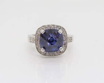 Op de afbeelding: Een zilveren ring met een grote, vierkante, donkerblauwe edelsteen omringd door een halo van kleinere, heldere edelstenen.