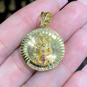 May include: A gold pendant featuring a detailed dragon design with a red gemstone in its mouth. The pendant is surrounded by a circle of clear cubic zirconia stones.