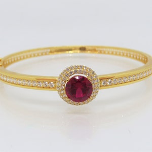 May include: A gold bangle bracelet featuring a red gemstone set in a halo of clear stones.