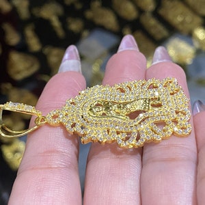 May include: A gold-colored pendant featuring a religious figure, encrusted with numerous small, clear gemstones. The pendant has an ornate, filigree-style border and a bail for attaching to a chain. The overall design is intricate and detailed.