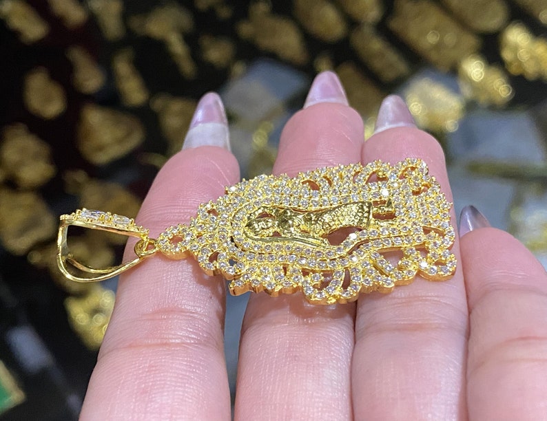 May include: A gold-colored pendant featuring a religious figure, encrusted with numerous small, clear gemstones. The pendant has an ornate, filigree-style border and a bail for attaching to a chain. The overall design is intricate and detailed.