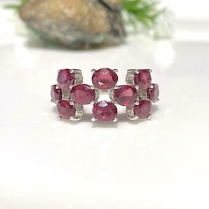 May include: A silver ring featuring multiple oval-cut, deep red gemstones. The gemstones are arranged in a cluster design, creating a bold and eye-catching piece of jewelry. The ring is set against a bright white background.