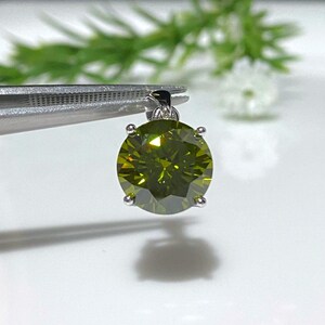 May include: A round green gemstone pendant with a silver setting. The pendant is held by tweezers.
