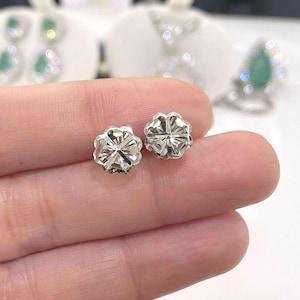 Puede incluir: Primer plano de dos peque&ntilde;os pendientes de bot&oacute;n plateados. Cada pendiente tiene un dise&ntilde;o floral con m&uacute;ltiples facetas, que reflejan la luz. Los pendientes est&aacute;n en la mano de una persona, con otras joyas visibles en el fondo.