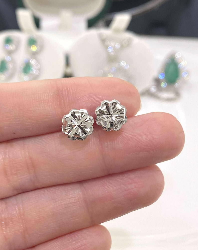 Puede incluir: Primer plano de dos peque&ntilde;os pendientes de bot&oacute;n plateados. Cada pendiente tiene un dise&ntilde;o floral con m&uacute;ltiples facetas, que reflejan la luz. Los pendientes est&aacute;n en la mano de una persona, con otras joyas visibles en el fondo.
