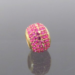 May include: A gold charm bead with a round shape and a pink crystal design. The bead is covered in small pink crystals.