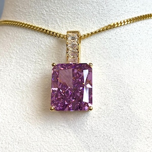 May include: A gold chain necklace with a large, rectangular, purple gemstone pendant. The pendant is surrounded by smaller, clear gemstones.