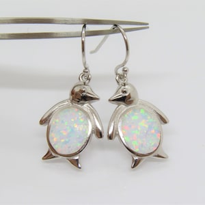 May include: A pair of silver-colored penguin earrings with white opal-like centers. The earrings have a hook closure and are displayed against a light background. The penguin design includes a beak, wings, and feet.
