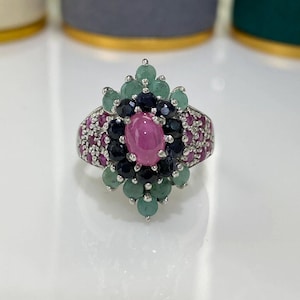 May include: A silver ring with a central oval pink gemstone, surrounded by smaller gemstones in shades of pink, green, and dark blue. The ring has a detailed, ornate design, showcasing a floral pattern.
