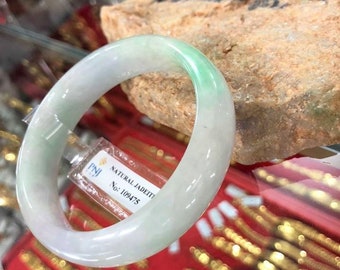 May include: A white jadeite bangle with a light green hue. The bangle is displayed on a rough stone with a tag that reads "NATURAL JADEITI No: 109475".