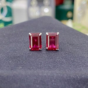 Könnte beinhalten: Ein Paar Ohrstecker mit rechteckigen, tiefroten Edelsteinen, die in silberfarbenem Metall gefasst sind. Die Ohrringe werden auf einer dunkelgrauen Oberfläche präsentiert, die ihre lebendige Farbe und ihr Design hervorhebt. Die Edelsteine haben eine polierte, reflektierende Oberfläche.