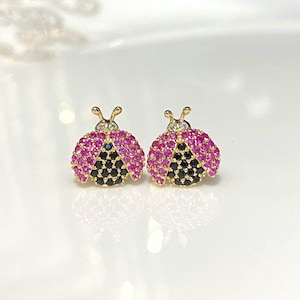 May include: A pair of gold ladybug stud earrings with pink and black cubic zirconia stones.