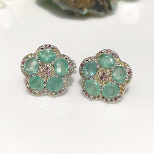 May include: A pair of flower-shaped stud earrings. Each earring features five oval-cut, light green gemstones surrounded by smaller pink and yellow stones. The earrings have a silver-colored metal setting.