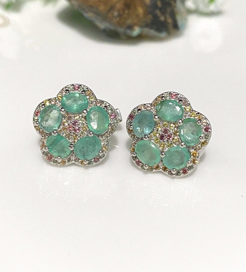 May include: A pair of flower-shaped stud earrings. Each earring features five oval-cut, light green gemstones surrounded by smaller pink and yellow stones. The earrings have a silver-colored metal setting.