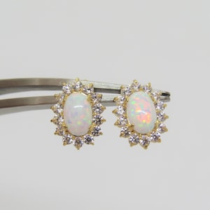 May include: A pair of gold stud earrings. Each earring has an oval, iridescent opal stone surrounded by a halo of small, clear gemstones. The earrings are held by a silver tweezer against a white background.
