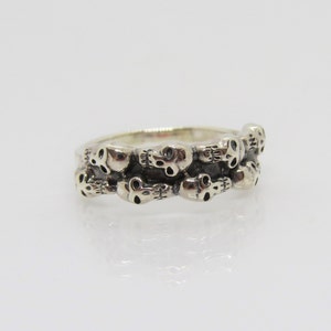 May include: A silver ring with a band of skulls. The skulls are arranged in a row around the band, and they are all facing the same direction.