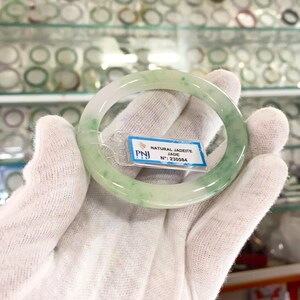 May include: A green jadeite bangle bracelet with a white background. The bracelet has a natural jadeite jade inscription and the number 230084.