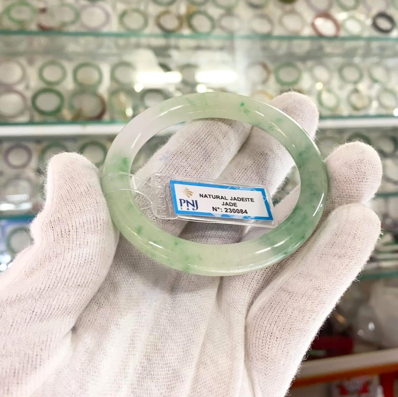 May include: A green jadeite bangle bracelet with a white background. The bracelet has a natural jadeite jade inscription and the number 230084.