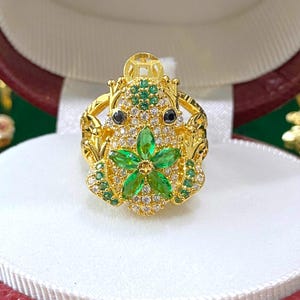 Peut inclure: Une bague en or avec un motif de grenouille verte. La grenouille tient une fleur verte dans ses pattes. La bague est sertie de pierres précieuses claires et vertes.