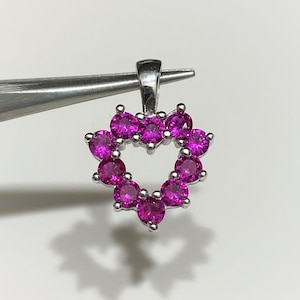 May include: A silver heart-shaped pendant with pink gemstones. The pendant is held by tweezers.