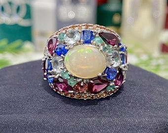 May include: A silver ring with a large oval opal centre stone, surrounded by an array of colourful gemstones. The ring features blue, purple, green, and clear stones, creating a vibrant design. The ring is set on a dark blue surface.