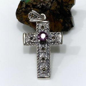 Pode incluir: Um pingente de cruz de prata com uma gema roxa no centro e detalhes de marcasita. A cruz é detalhada com uma borda de corda e desenhos em forma de estrela.