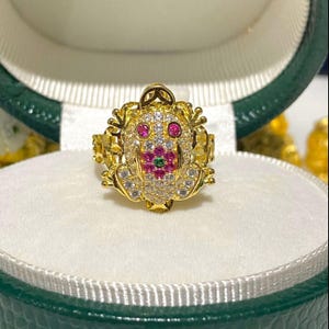 以下が含まれることがあります： 詳細なデザインの金色のカエルリング。カエルの目は赤く、ピンクと緑のアクセントの花が顔を飾っています。リングには、小さな透明な宝石がちりばめられています。