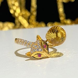 May include: A gold ring with a fox design and a clear yellow gemstone. The fox has a red ruby on its nose and white cubic zirconia stones for eyes.