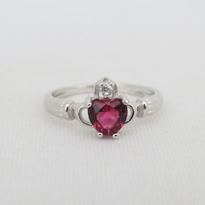 May include: A silver ring with a red heart-shaped gemstone and a smaller clear gemstone on top. The ring is a Claddagh ring, a traditional Irish design.