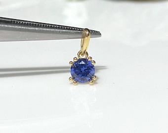 Pode incluir: Um pingente de ouro com uma gema azul engastada em um bezel redondo. O pingente está sendo segurado por uma pinça.