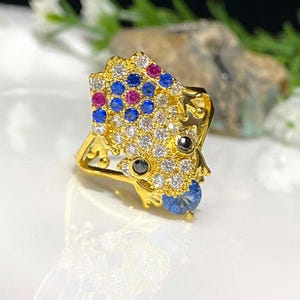 Könnte beinhalten: Ein goldener Ring mit einem Froschdesign. Der Frosch ist mit klaren, blauen und rosa Edelsteinen verziert. Der Ring ist ein Statement-Piece und wäre ein tolles Geschenk.