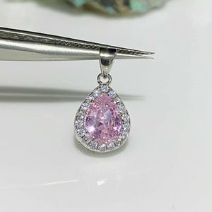 May include: A teardrop-shaped pendant with a large, light pink center stone surrounded by a halo of smaller, clear stones. The pendant is suspended from a silver bail. The jewelry is held by a pair of tweezers.