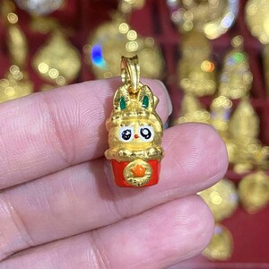 May include: A gold-colored pendant featuring a cartoon bunny character with green accents and black eyes, atop a red base with a star emblem. The pendant is held in a hand, with other gold pendants visible in the background.