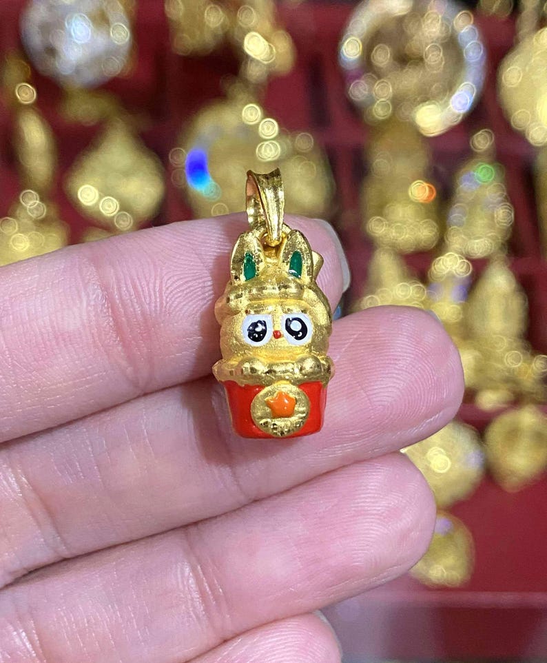 May include: A gold-colored pendant featuring a cartoon bunny character with green accents and black eyes, atop a red base with a star emblem. The pendant is held in a hand, with other gold pendants visible in the background.