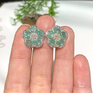 May include: A pair of flower-shaped stud earrings. Each earring features five oval-cut, light green gemstones surrounded by smaller pink and yellow stones, set in a silver-colored metal. The earrings are displayed against a neutral background.