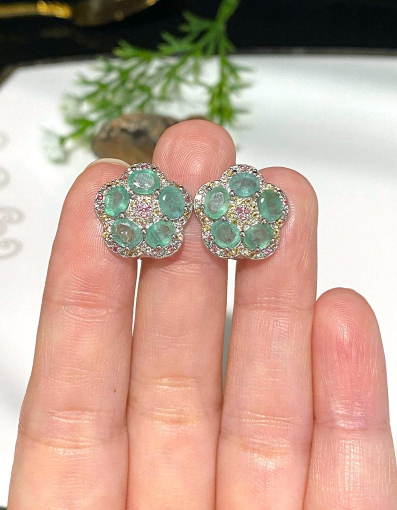 May include: A pair of flower-shaped stud earrings. Each earring features five oval-cut, light green gemstones surrounded by smaller pink and yellow stones, set in a silver-colored metal. The earrings are displayed against a neutral background.