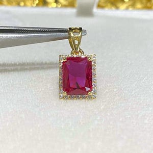 Pode incluir: Pingente de ouro com uma pedra preciosa retangular, vermelho escuro no centro. A pedra preciosa é rodeada por um halo de pedras menores e transparentes. O pingente é segurado por uma pinça contra um fundo branco macio.